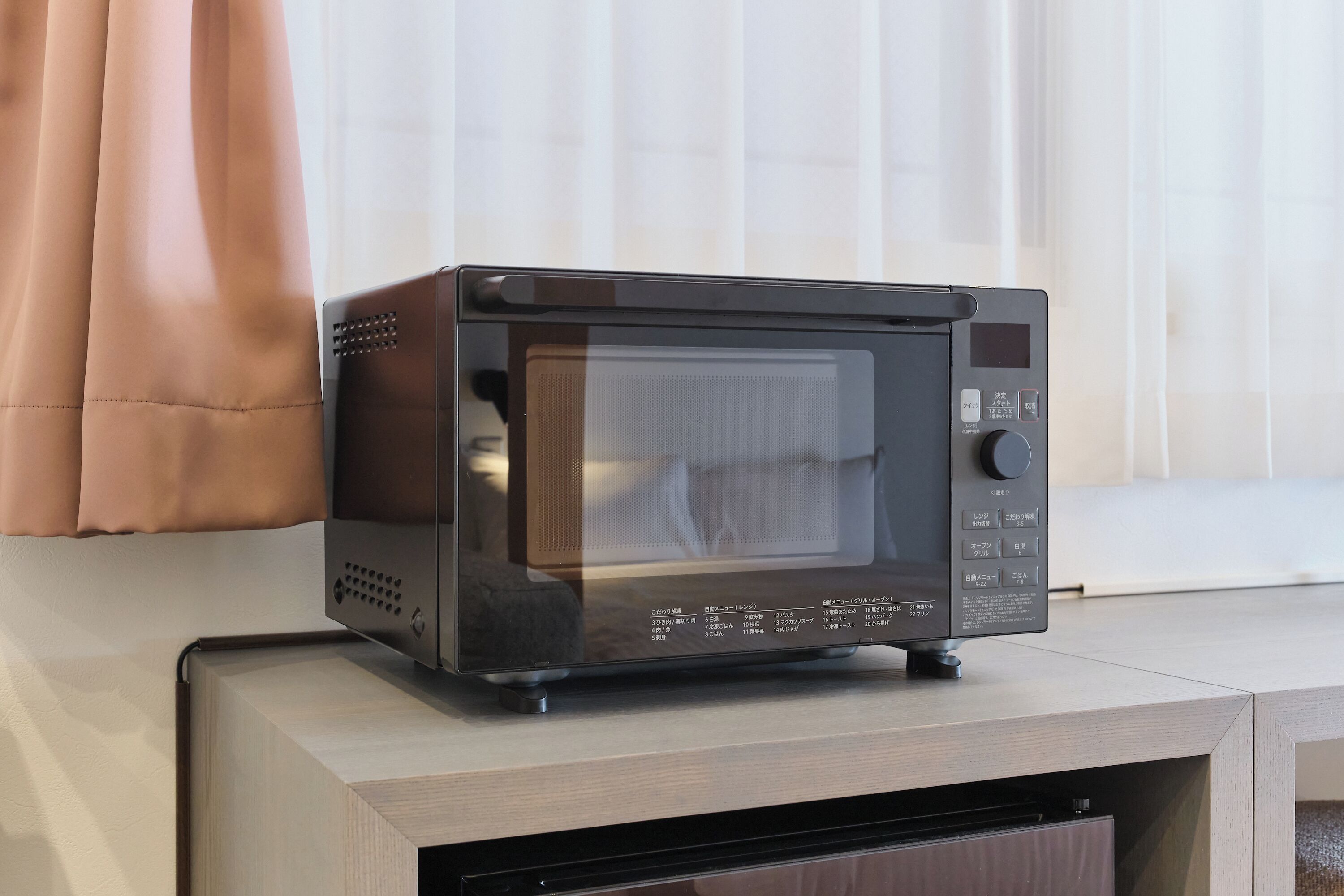 A microwave on a wooden cabinet in a room with a window and curtains.