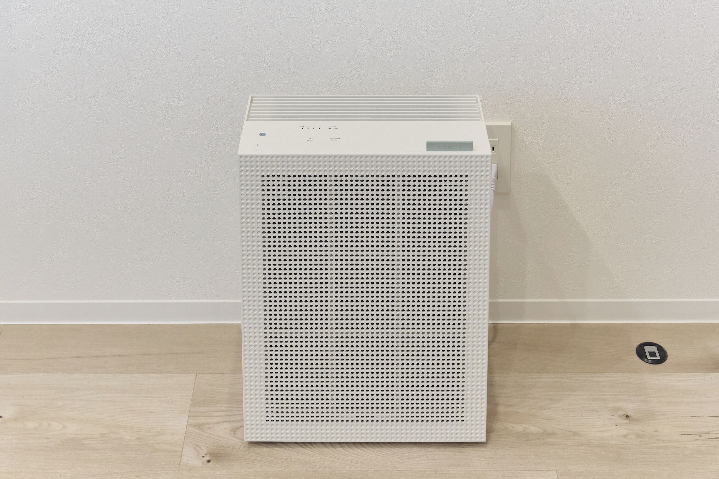A white, rectangular, freestanding heater with a perforated front panel, placed on a wooden floor against a plain wall.