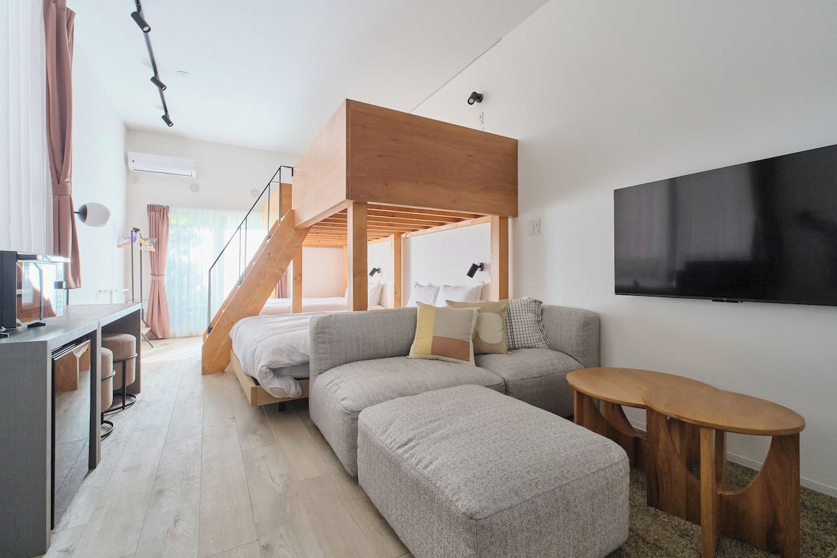 A modern living space with a loft bed, a sofa, a small table, and a TV mounted on the wall.