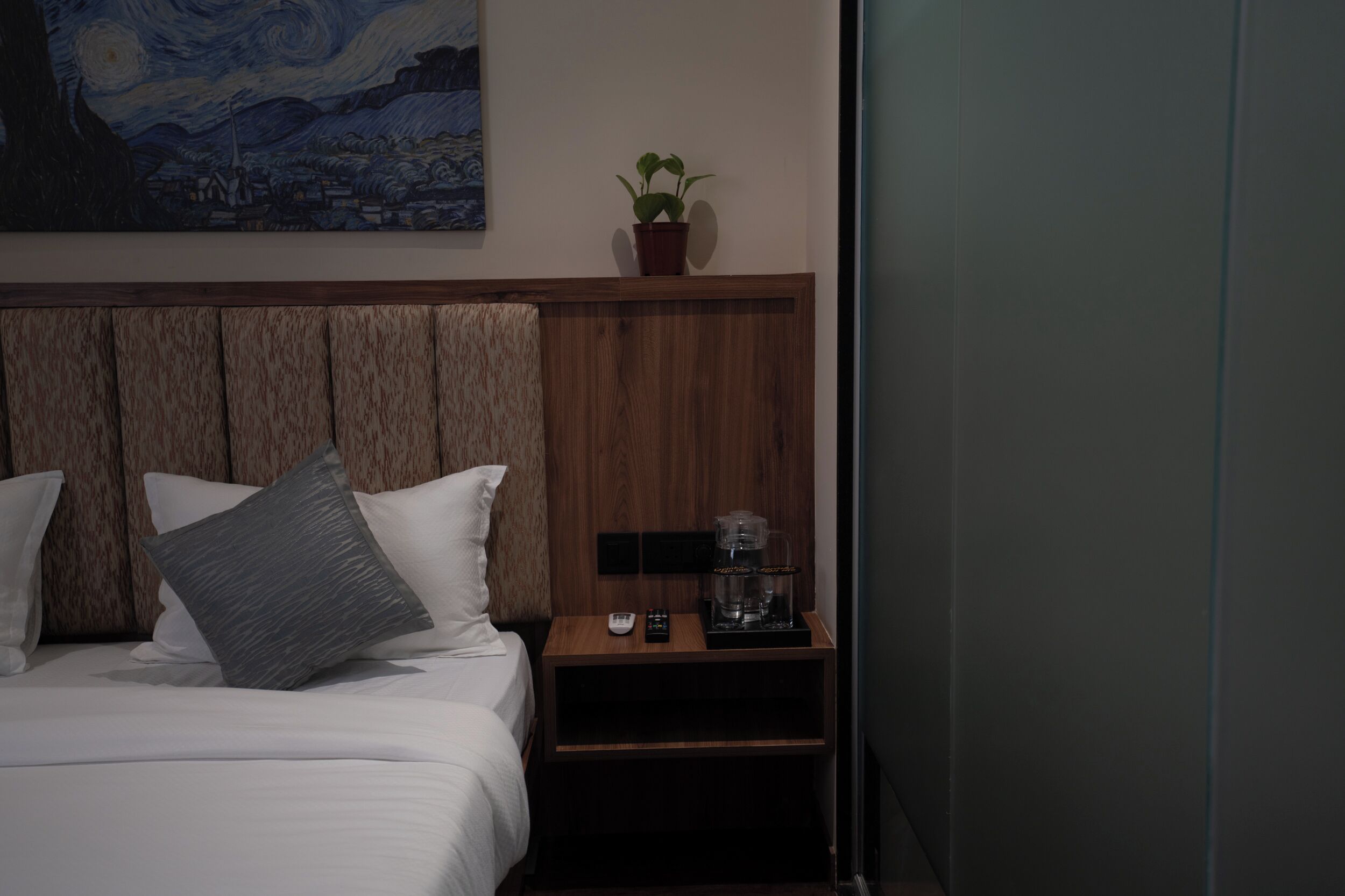 Een hotelkamer met een houten hoofdeinde, een bed met wit beddengoed, een nachtkastje met een koffiezetapparaat, een afstandsbediening en een kleine potplant.