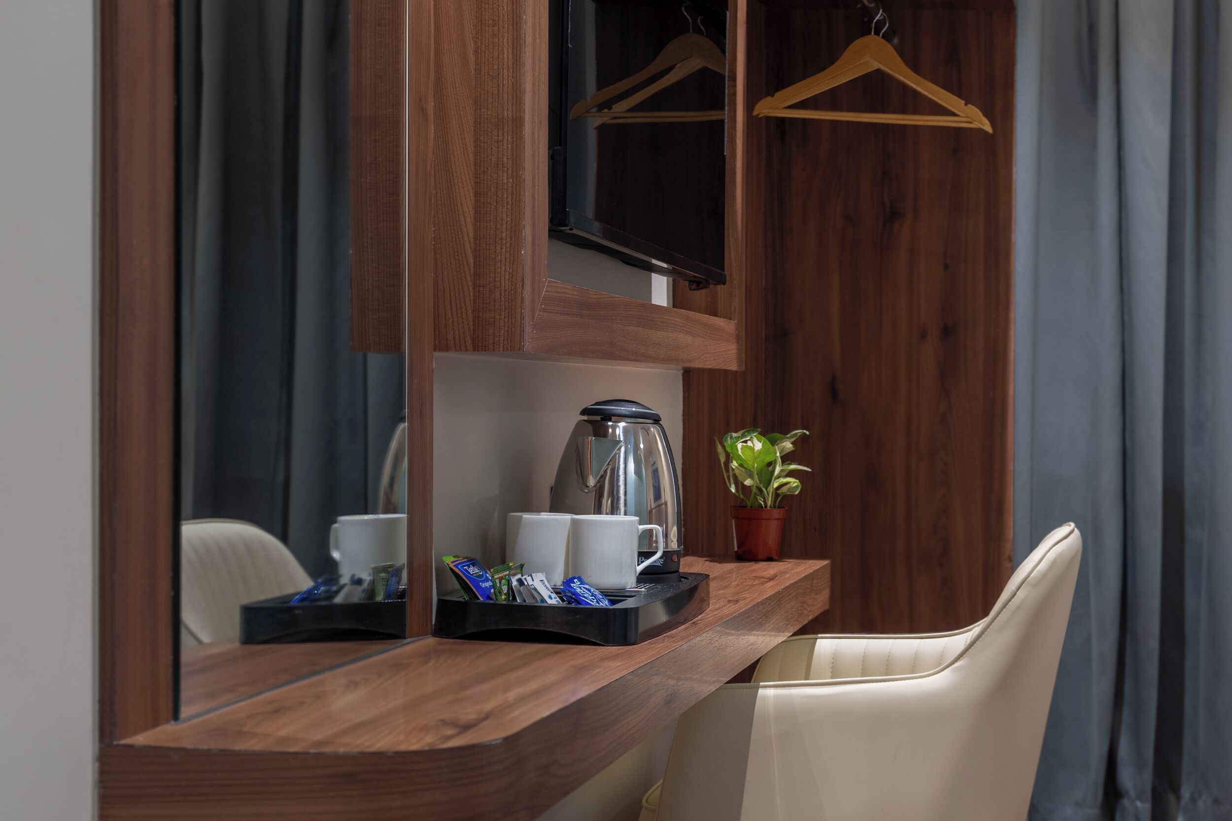 Una mesa de madera con una cafetera, tazas y una bandeja con bocadillos. Hay una planta en maceta sobre la mesa, y se ve una silla.