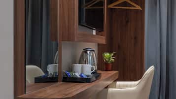 Una mesa de madera con una cafetera, tazas y una bandeja con bocadillos. Hay una planta en maceta sobre la mesa, y se ve una silla.
