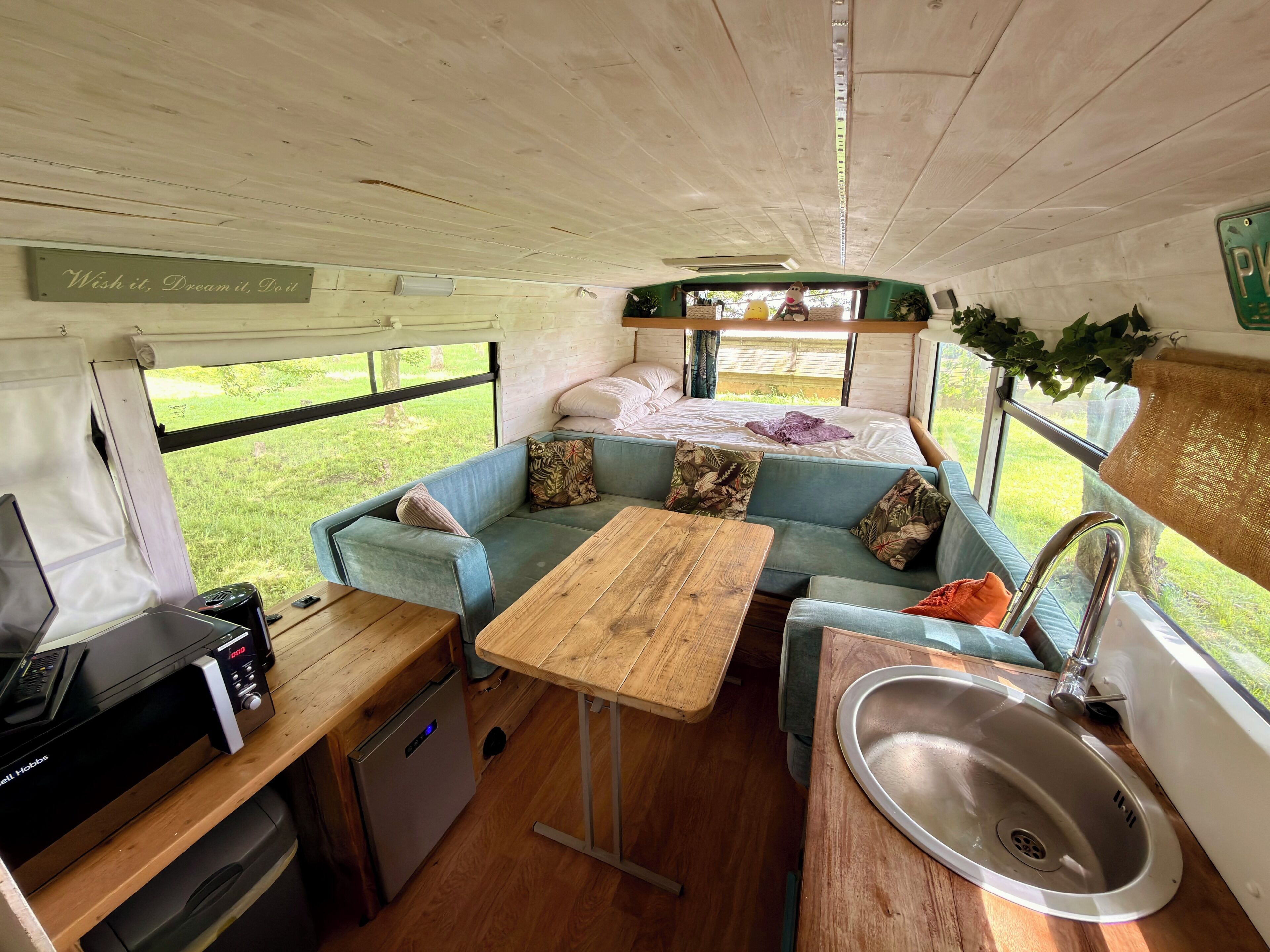 A compact RV interior with a kitchenette, a dining area, a bed, and a window providing a view of the outdoors.
