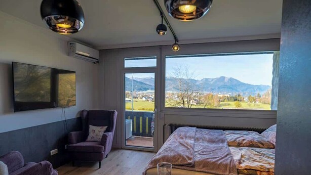 Ett sovrum med en säng, en stol, ett bord, en tv och ett stort fönster med utsikt över bergen.
