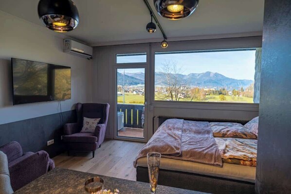 Ein Schlafzimmer mit Bett, Stuhl, Tisch, Fernseher und einem großen Fenster mit Blick auf die Berge.