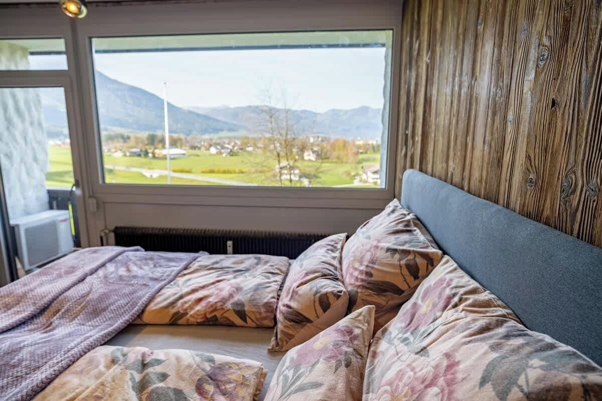 A bed with floral bedding, a view of a landscape through a window, and a wooden wall.