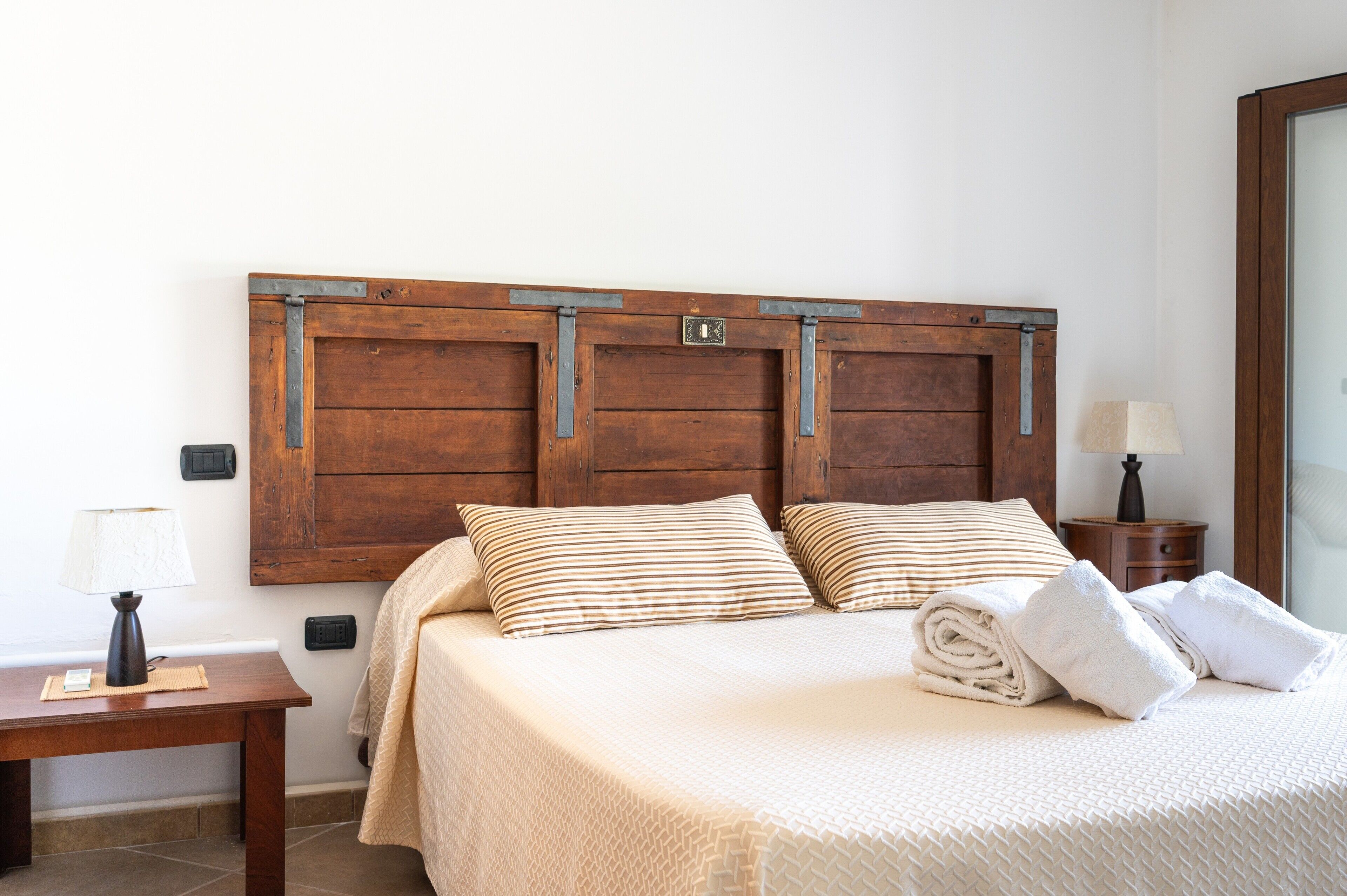 Un letto rifatto con cuscini a righe, una testiera in legno, un comodino con una lampada e un tavolino notte con una lampada.