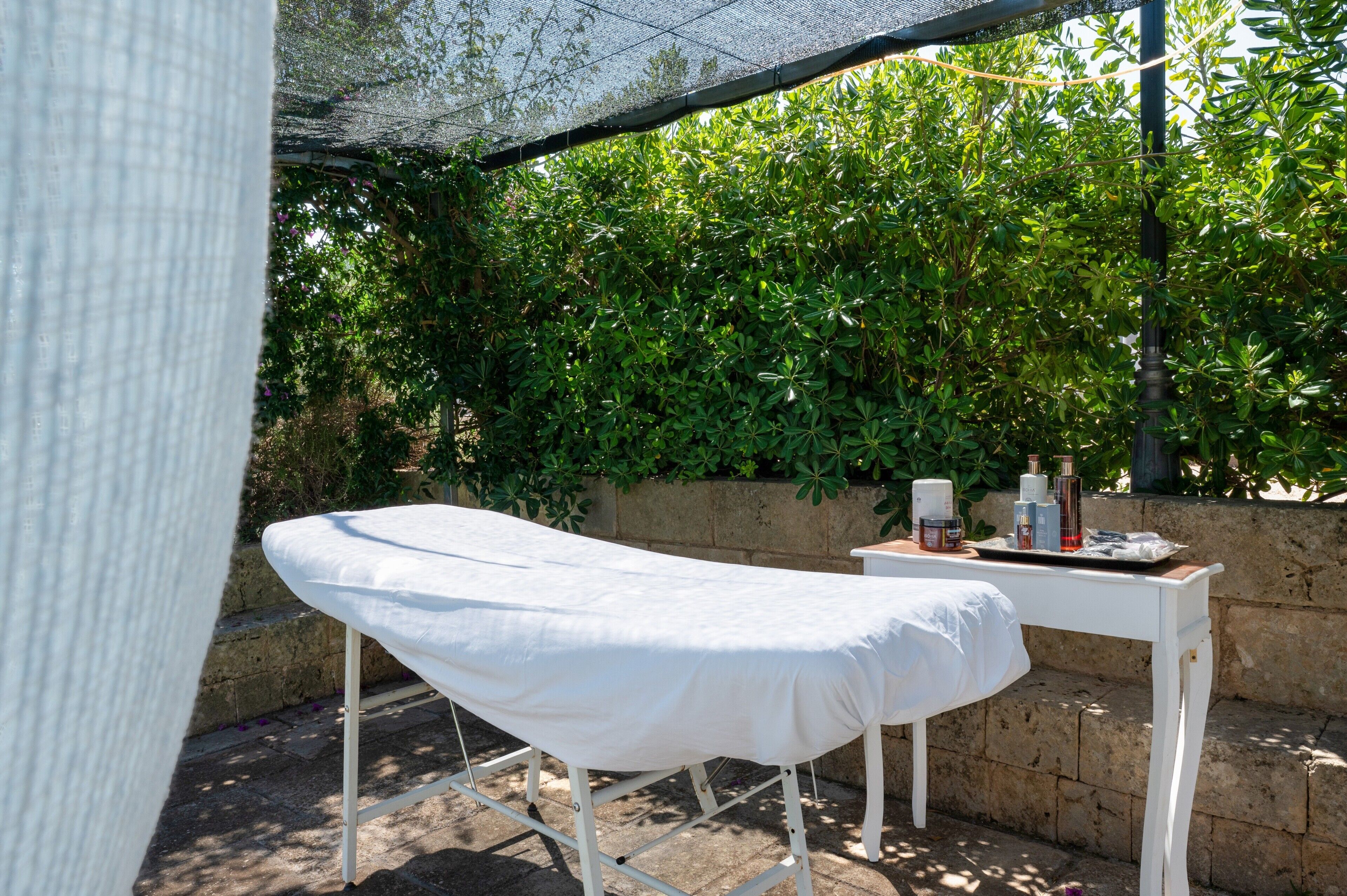 A treatment table with a white sheet, a small table with bottles, and a stone wall in a shaded outdoor area.