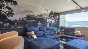 Ett modernt vardagsrum med en stor hörnsoffa, ett soffbord i glas och utsikt över ett slott genom ett stort fönster.