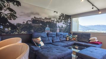 Ein modernes Wohnzimmer mit einer großen Eckcouch, einem Glastisch und Blick auf eine Burg durch ein großes Fenster.