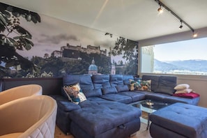 A modern living room with a large sectional sofa, a glass coffee table, and a view of a castle through a large window. - Premium Lake view Suite | Adults only (Strobl)