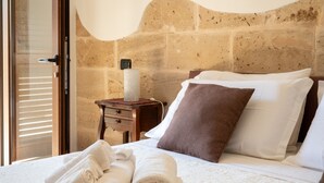 Una habitación de hotel con pared de piedra, cama con ropa blanca, almohada marrón y una toalla enrollada sobre la cama.