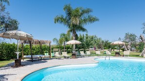 Zona de piscina con sillones de descanso y sombrillas, rodeada de follaje y palmeras.