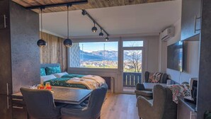 Un dormitorio moderno con una cama grande, una mesa de comedor y sillas, un televisor, y vistas a un pueblo y montañas.