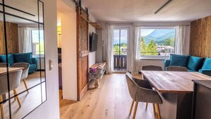 Una habitación moderna con suelo de madera, una mesa de comedor con sillas, un sofá, un televisor montado en una pared de madera y un ventanal con vistas a montañas.