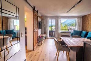 Ein modernes Zimmer mit Holzboden, einem Esstisch mit Stühlen, einer Couch, einem an einer Holzwand montierten Fernseher und einem großen Fenster mit Bergblick.