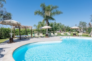 Piscina com espreguiçadeiras e guarda-sóis, cercada por palmeiras e vegetação.