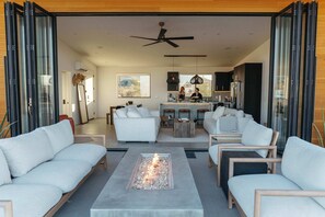 Ein modernes Wohnzimmer mit Kamin, weißen Sofas und einem Essbereich, im Hintergrund ist eine Küche zu sehen.