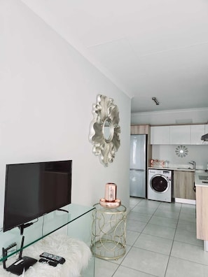 Ein modernes Wohnzimmer mit einem Flachbildfernseher, einem Glastisch, einem flauschigen weißen Teppich, einem dekorativen Spiegel und einer Küchenzeile mit einer Waschmaschine.