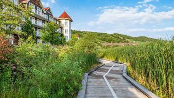 A wooden boardwalk leading to a building with a red roof, surrounded by lush greenery and hills in the background.