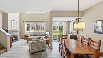 A spacious living room with a fireplace, a dining area with a wooden table and chairs, and a large window offering a view of the outdoors.
