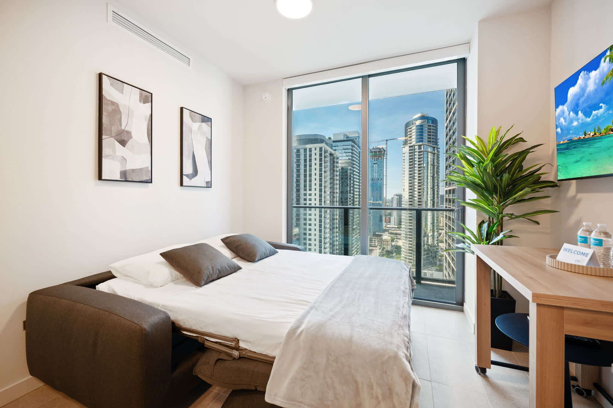 A modern hotel room with a bed, a desk, a large window with a city view, and two abstract paintings on the wall.