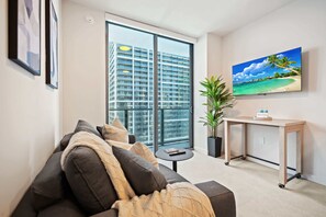 A modern living room with a sofa, a small table, a potted plant, a wall-mounted TV, and a large window with a city view. - 501 FIRST LUXURY 1BR+DEN (Miami)