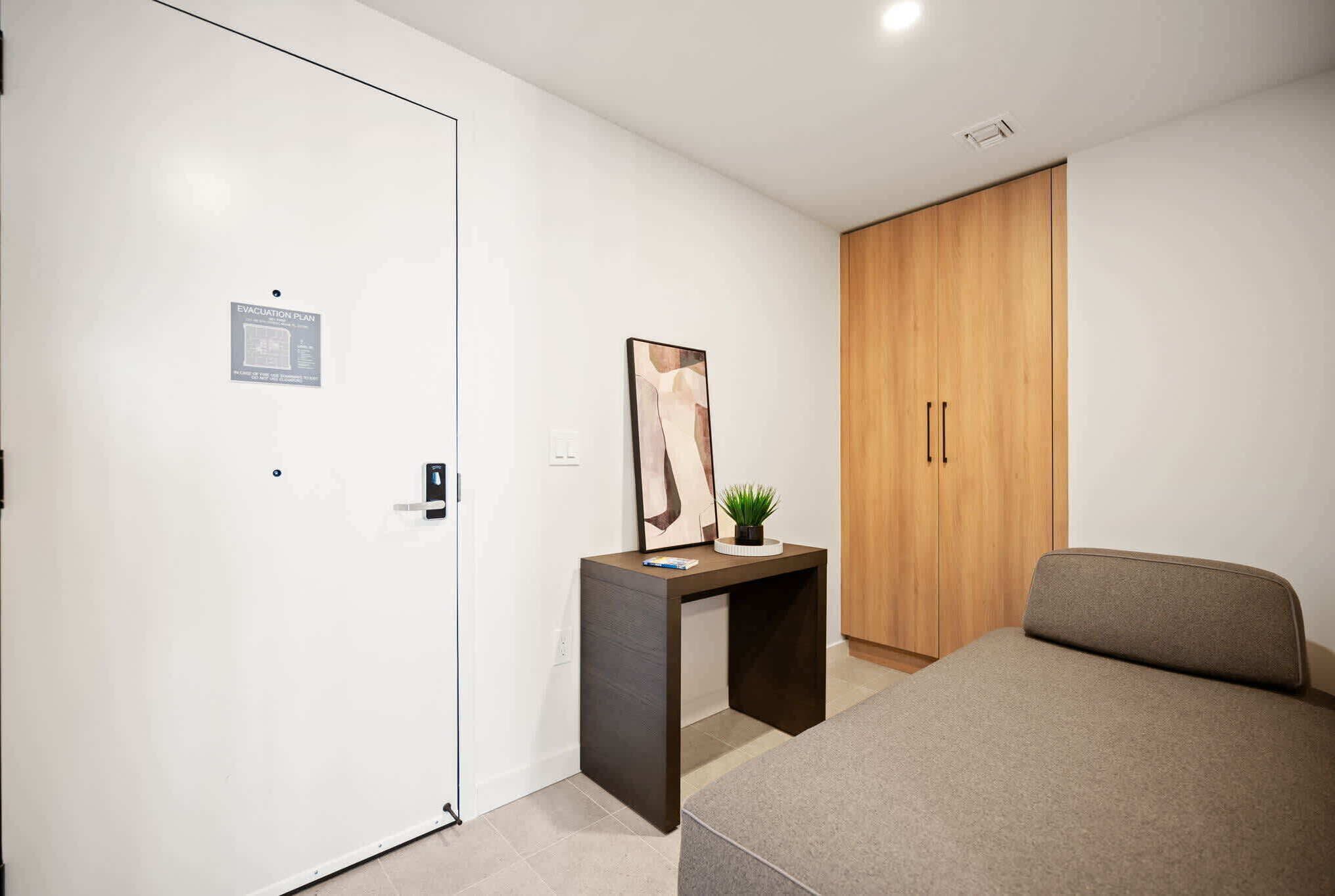 A bedroom with a bed, a desk, a wardrobe, and a door with an evacuation plan sign.