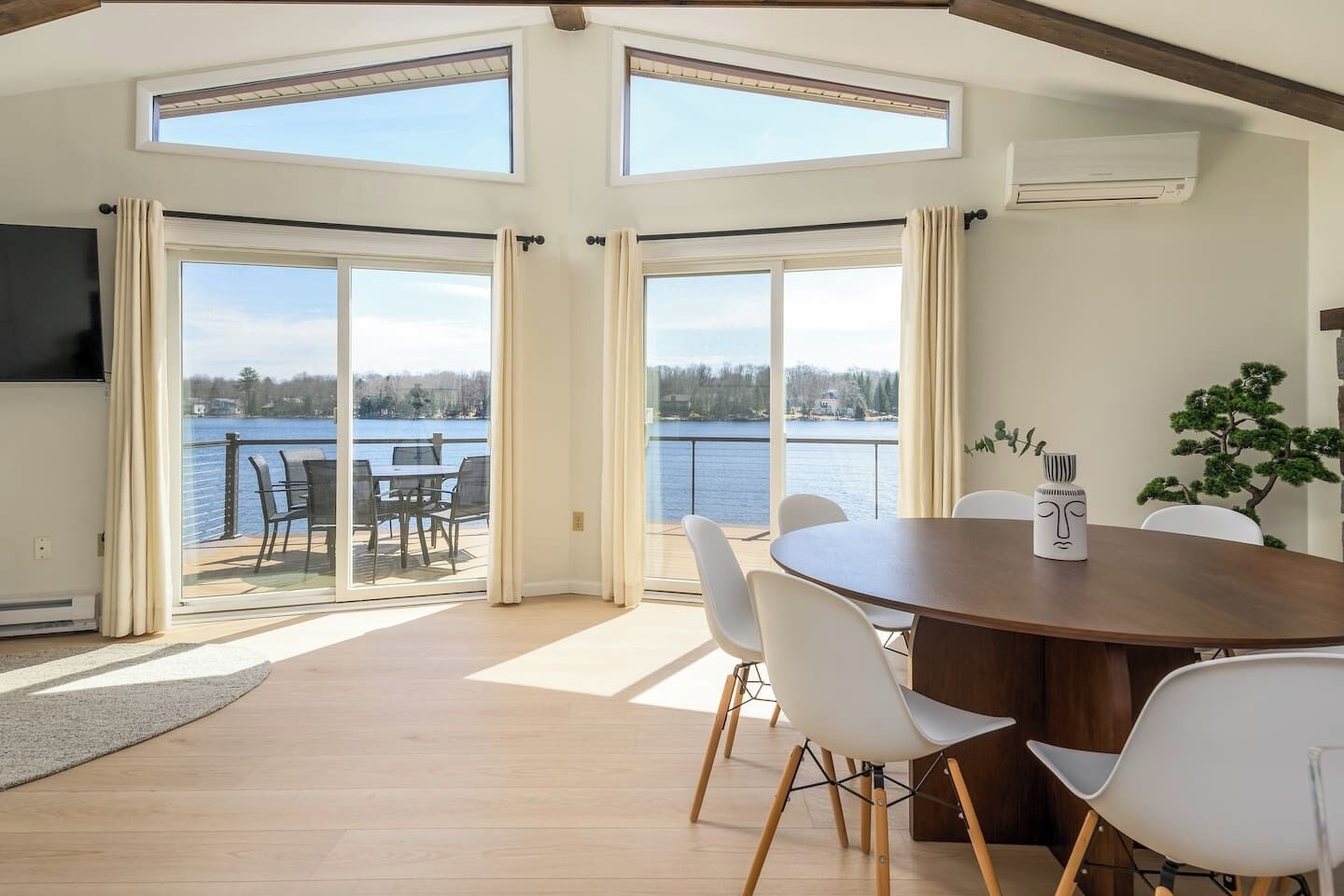 Una habitación con una mesa grande, sillas blancas, un televisor y vistas a un lago.