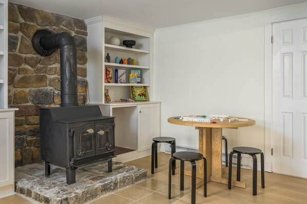 A stone wall, a black wood-burning stove, a white shelf with books, and a round wooden table with stools.