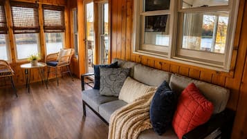 A cozy cabin interior with a wooden floor, a sofa with patterned cushions, a table with chairs, and large windows overlooking a body of water.