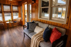Een gezellig blokhutinterieur met een houten vloer, een bank met gestreepte kussens, een tafel met stoelen, en grote ramen met uitzicht op een wateroppervlak.