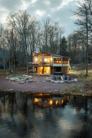 Een chalet met twee verdiepingen, een terras, een vuurplaats en een vijver ervoor.