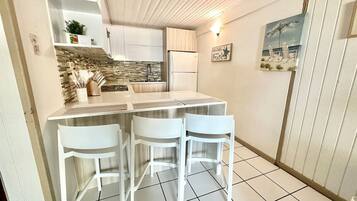 A compact kitchen with a central island, white bar stools, a refrigerator, and a tiled floor.