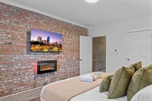 Una camera d'albergo con una parete in mattoni, una TV a schermo piatto sopra un caminetto e un letto con cuscini, e una porta.