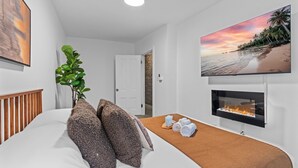Un dormitorio con cama, una planta, chimenea y televisor de pared que muestra una escena de playa.