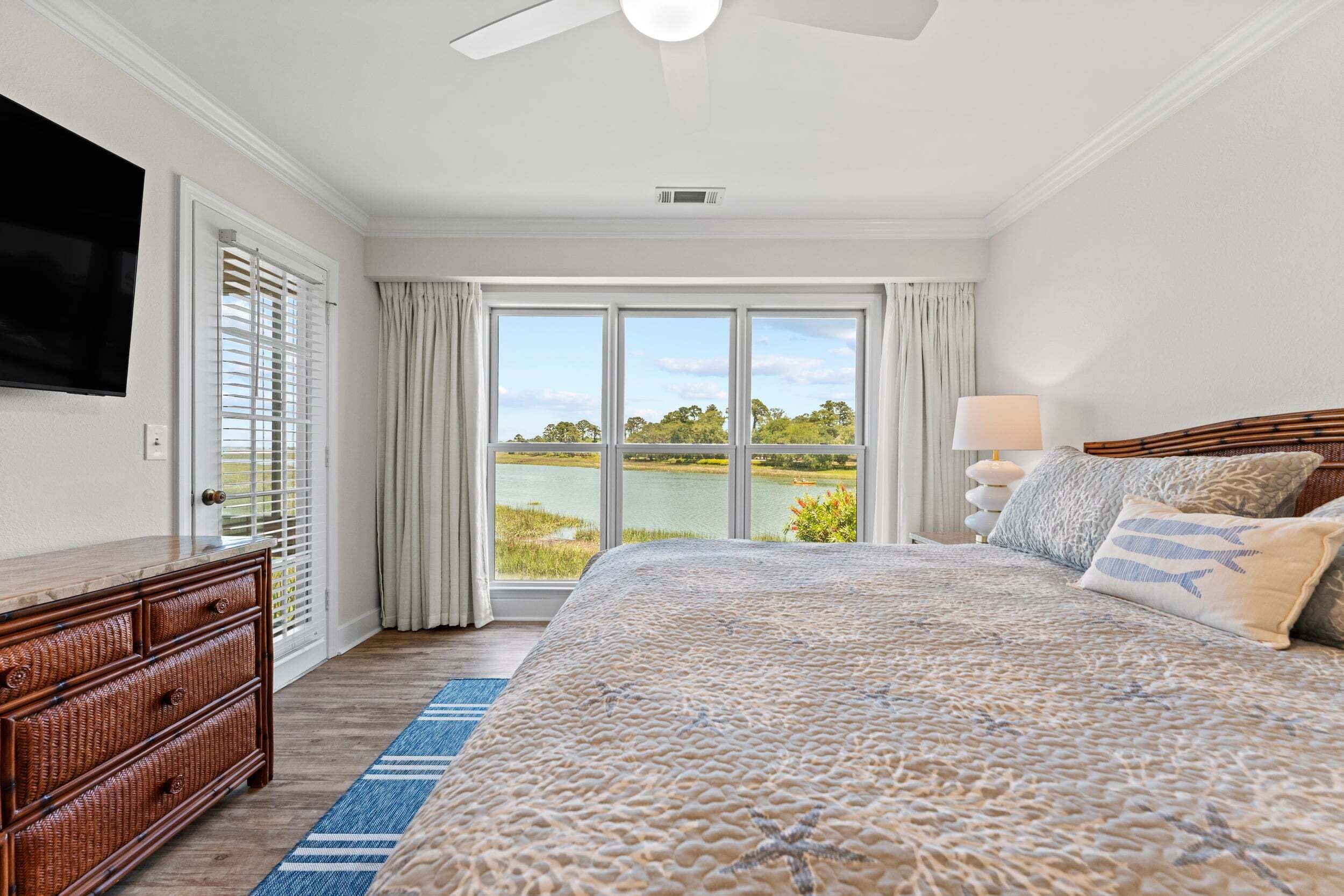 A bedroom with a large bed, a television on the wall, a dresser, and a view of a lake.