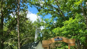 Un pont en bois mène à une petite chapelle blanche surmontée d’un clocher, le tout étant entouré d’une végétation luxuriante.
