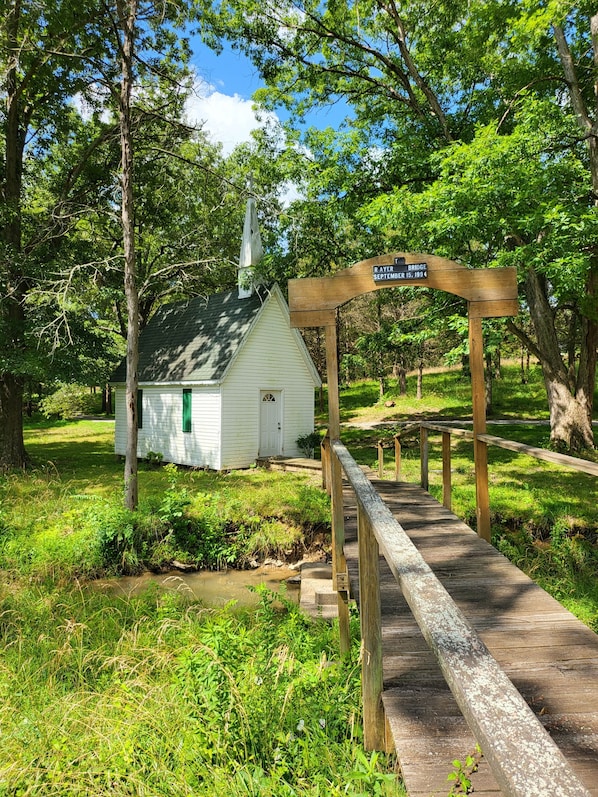 A wooden bridge leading to a small white chapel with a steeple, surrounded by lush greenery.