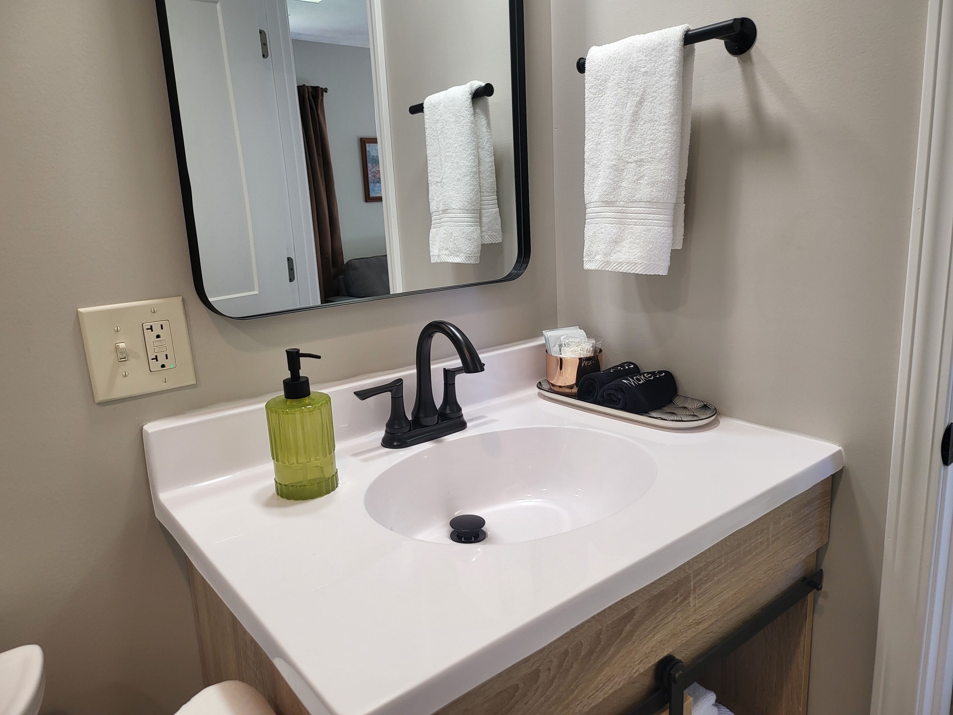 A bathroom with a white sink, black faucet, a mirror, a towel rack with a white towel, a green soap dispenser, and a small tray with a towel and a bottle.