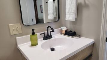 A bathroom with a white sink, black faucet, a mirror, a towel rack with a white towel, a green soap dispenser, and a small tray with a towel and a bottle.
