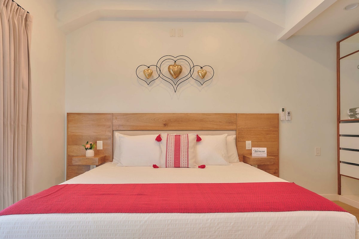 Habitación de hotel con cabecera de madera, colcha roja y decoración en forma de corazón en la pared.