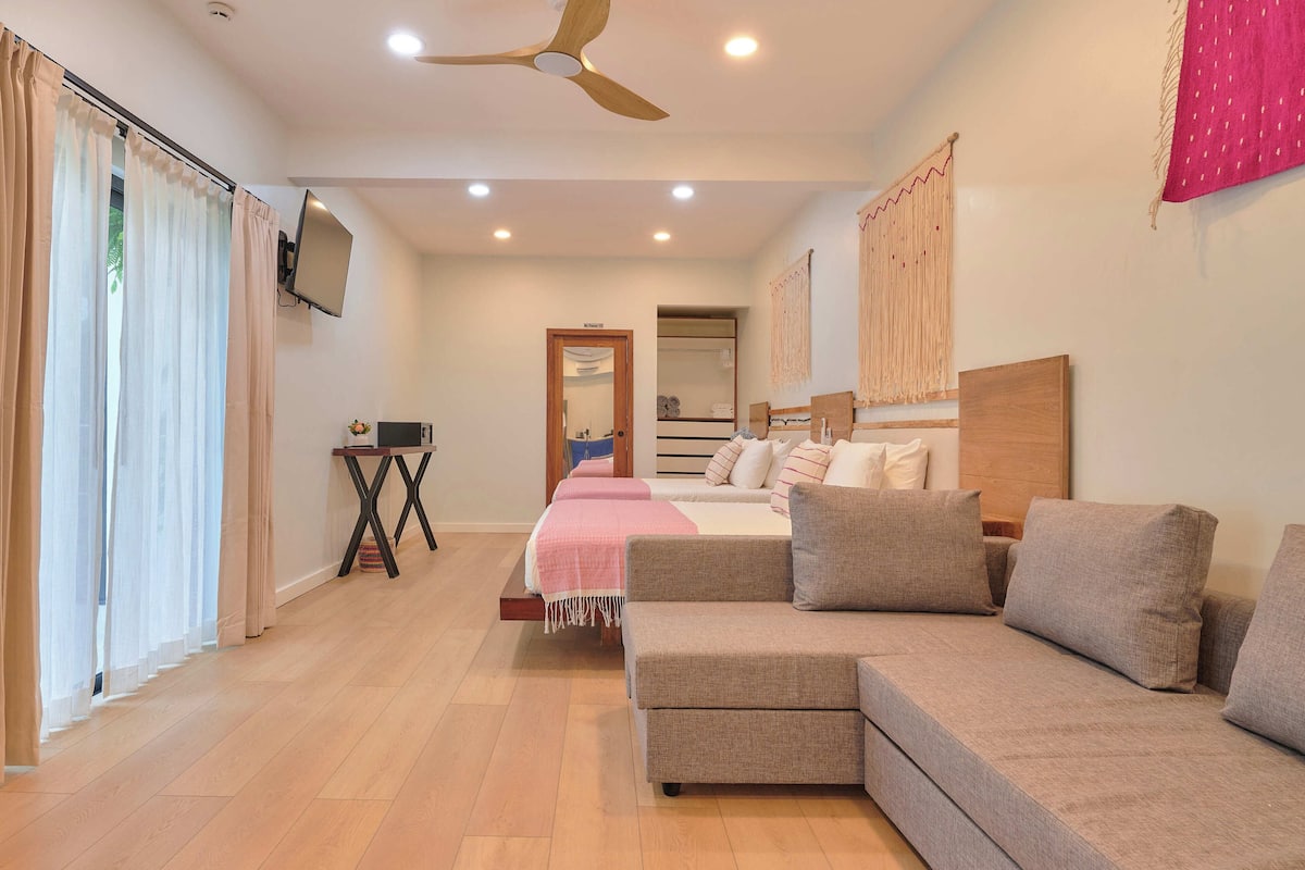 Habitación de hotel con sofá, cama con ropa de cama rosa, televisor montado en la pared y ventilador de techo.