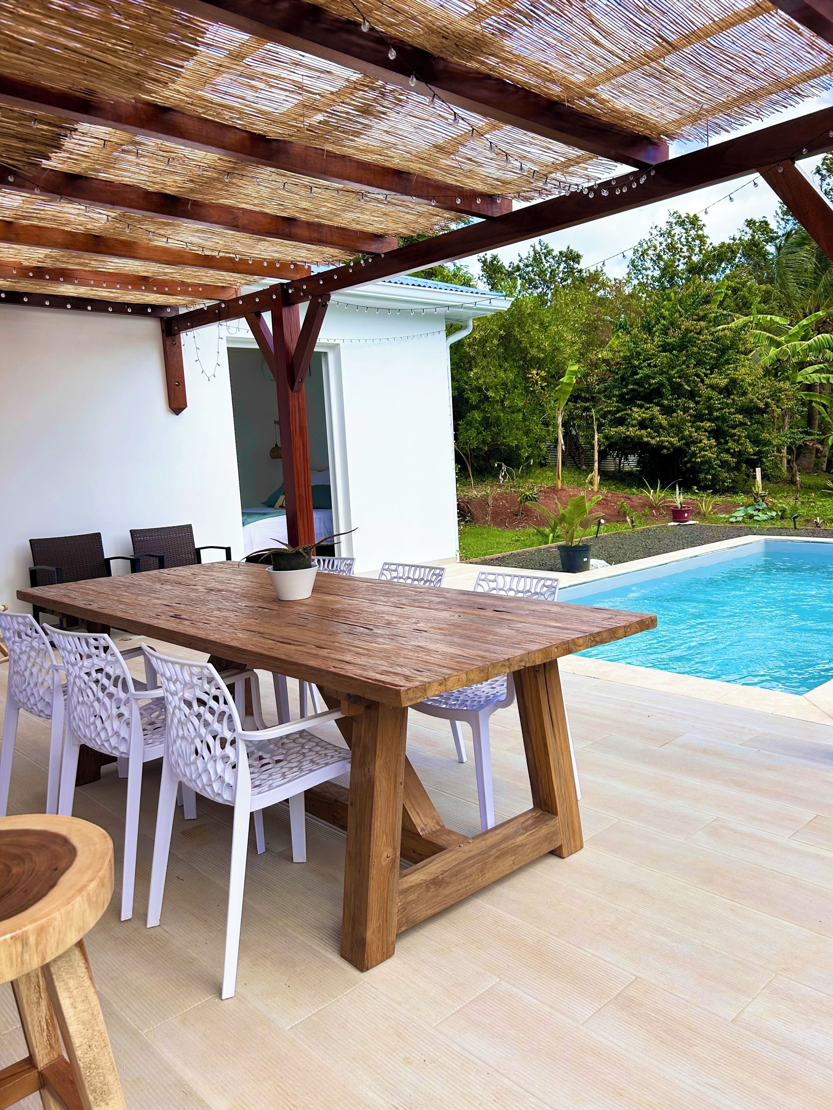 An outdoor patio with a wooden table and white chairs, a pool, and a covered area with a thatched roof.