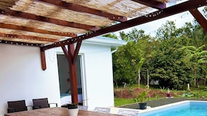 Eine Außenpatio mit Holztisch und weißen Stühlen, einem Pool und einem überdachten Bereich mit einem Reetdach.