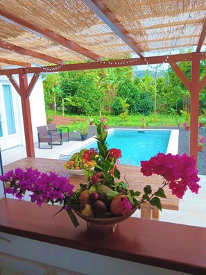 A covered outdoor area with a pool, a table with fruit and flowers, and a view of lush greenery. - Casa Helena – Brand-New Villa – Private Pool &Tropical Garden – 6 P – Martinique (Saint-Esprit)