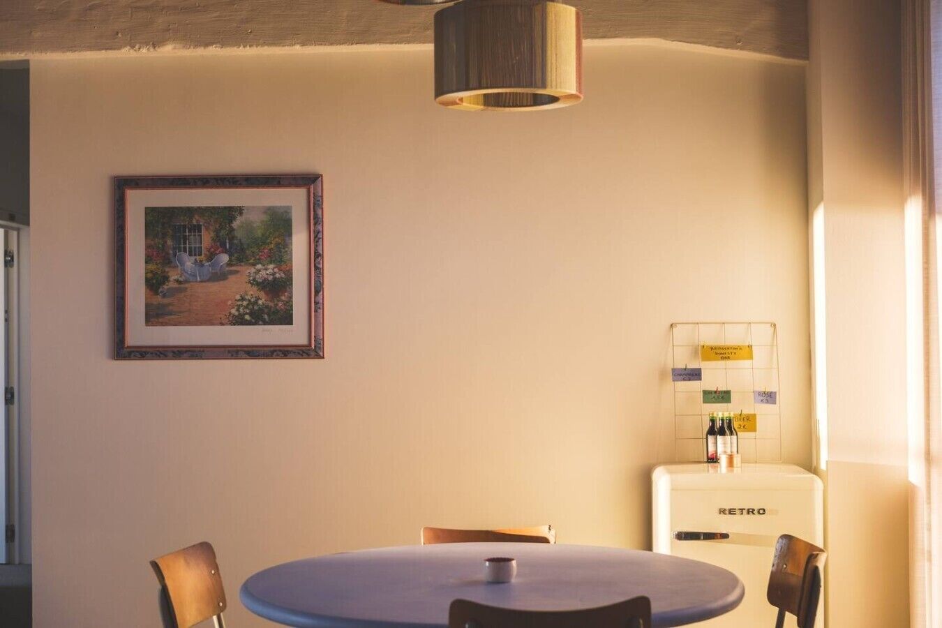 Een eethoek met een ronde tafel, stoelen, een lijst met een schilderij, een waterdispenser en een hanglamp.