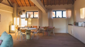 Una habitación amplia con techo de madera, comedor con mesa y sillas, kitchenette y ventanas grandes que dejan entrar luz natural.