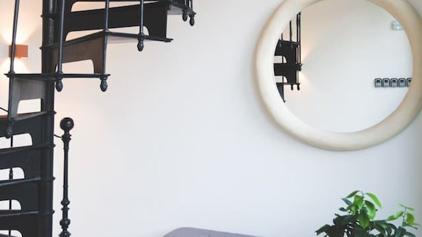 Una escalera de caracol, un espejo redondo, un banco y una planta en maceta en una habitación.