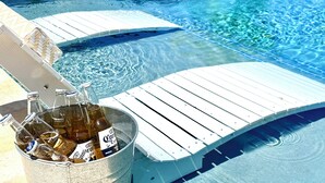 Uma área à beira da piscina com uma espreguiçadeira de madeira, um balde com garrafas de cerveja e um caiaque vermelho ao fundo.